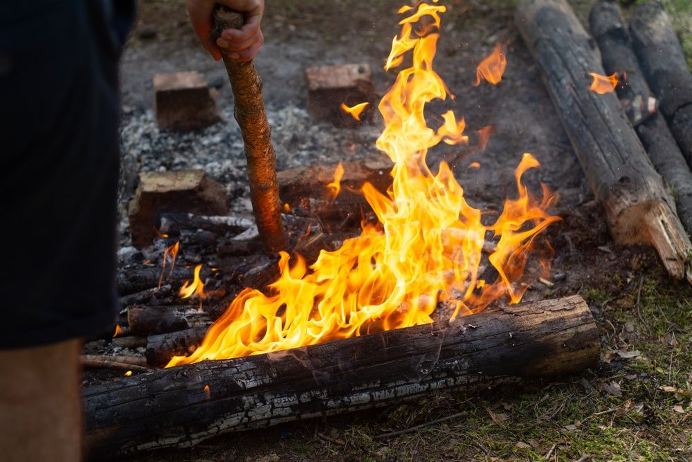 A person uses a wooden stick to stir burning logs in an outdoor campfire.