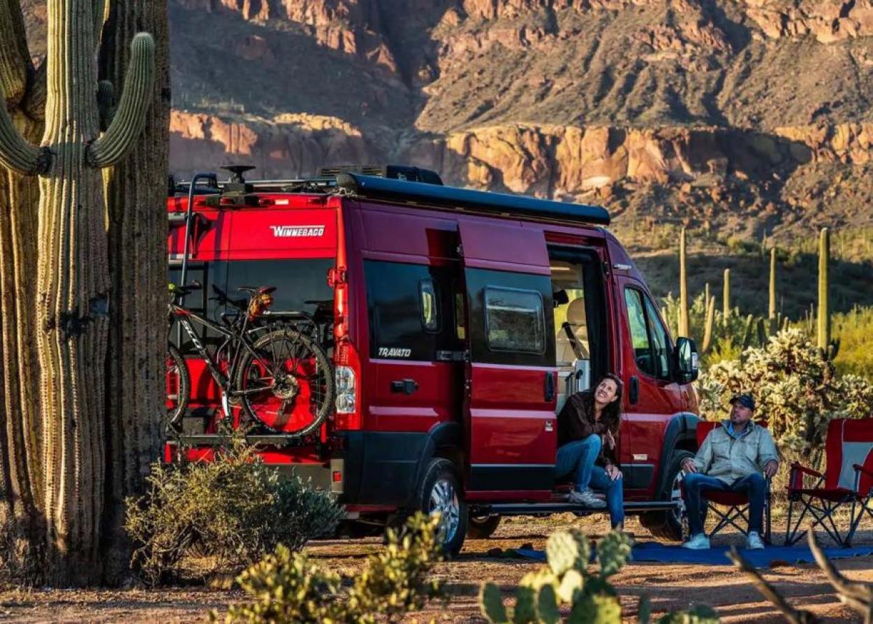 Red 2023 Winnebago Travato parked in a desert by a cactus, with a woman sitting in the doorway and a man in a camping chair outside of the van