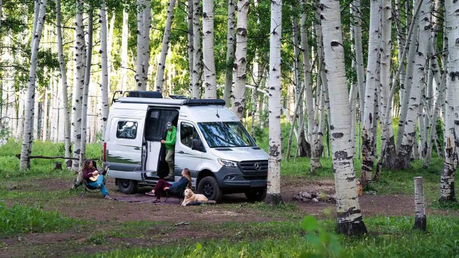 Family included one man, two women, and a yellow lab sitting outside a parked Winnebago Revel campervan in a wooded area