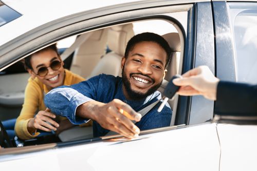 A smiling man and a woman sit in a car, as the man accepts a key from someone standing outside 