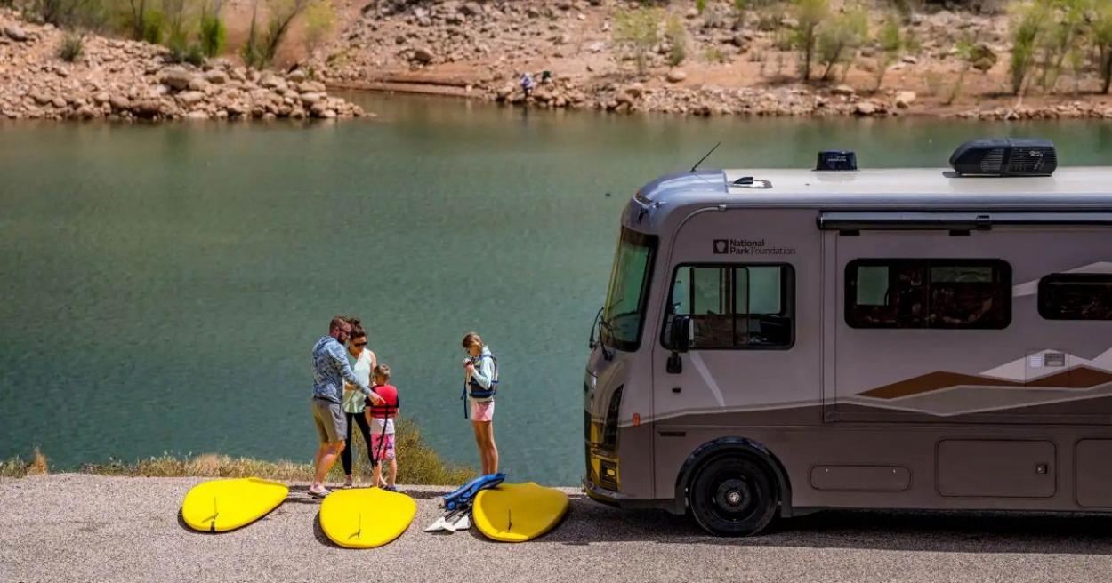 Family loading their paddle boards on to the river with their Winnebago Class A RV parked nearby