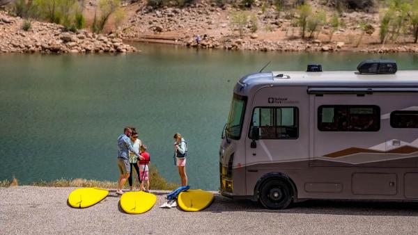 Family loading their paddle boards on to the river with their Winnebago Class A RV parked nearby