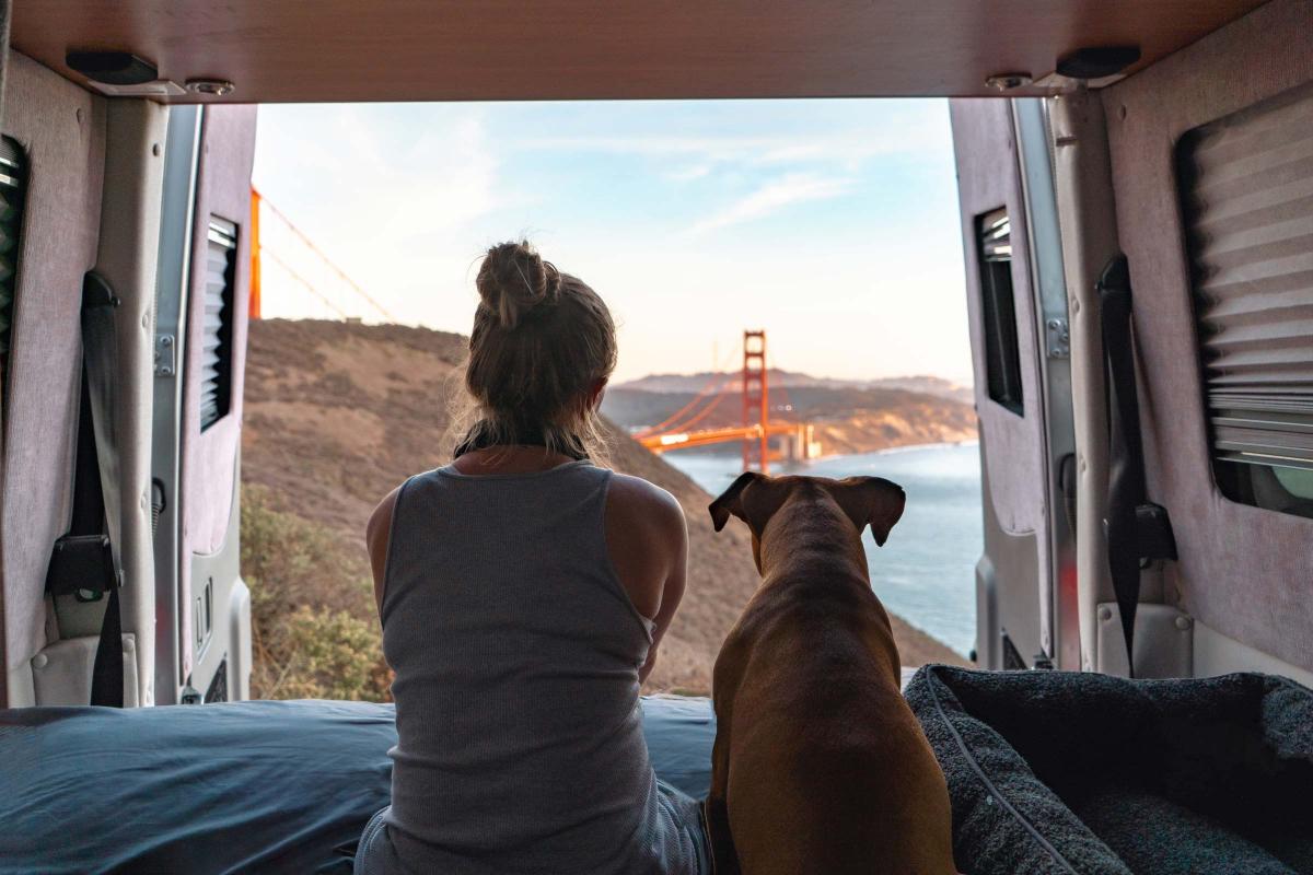 Woman and her dog sitting at the back of their campervan, which has its doors open. They are facing away and you can see the Golden Gate Bridge in the distance