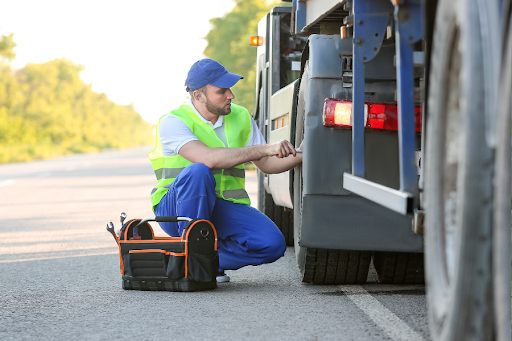 Mechanic in safety vest kneeling by a commercial trailer tire on the roadside, inspecting or repairing it with a tool bag placed nearby.