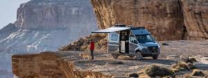 Man and dog standing outside a Winnebago Revel parked on a rocky outcrop, with the campervan&#039;s awning outstretched