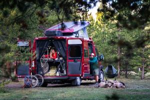 Couple in the rear of a red Winnebago campervan, setting up camp in a forested area