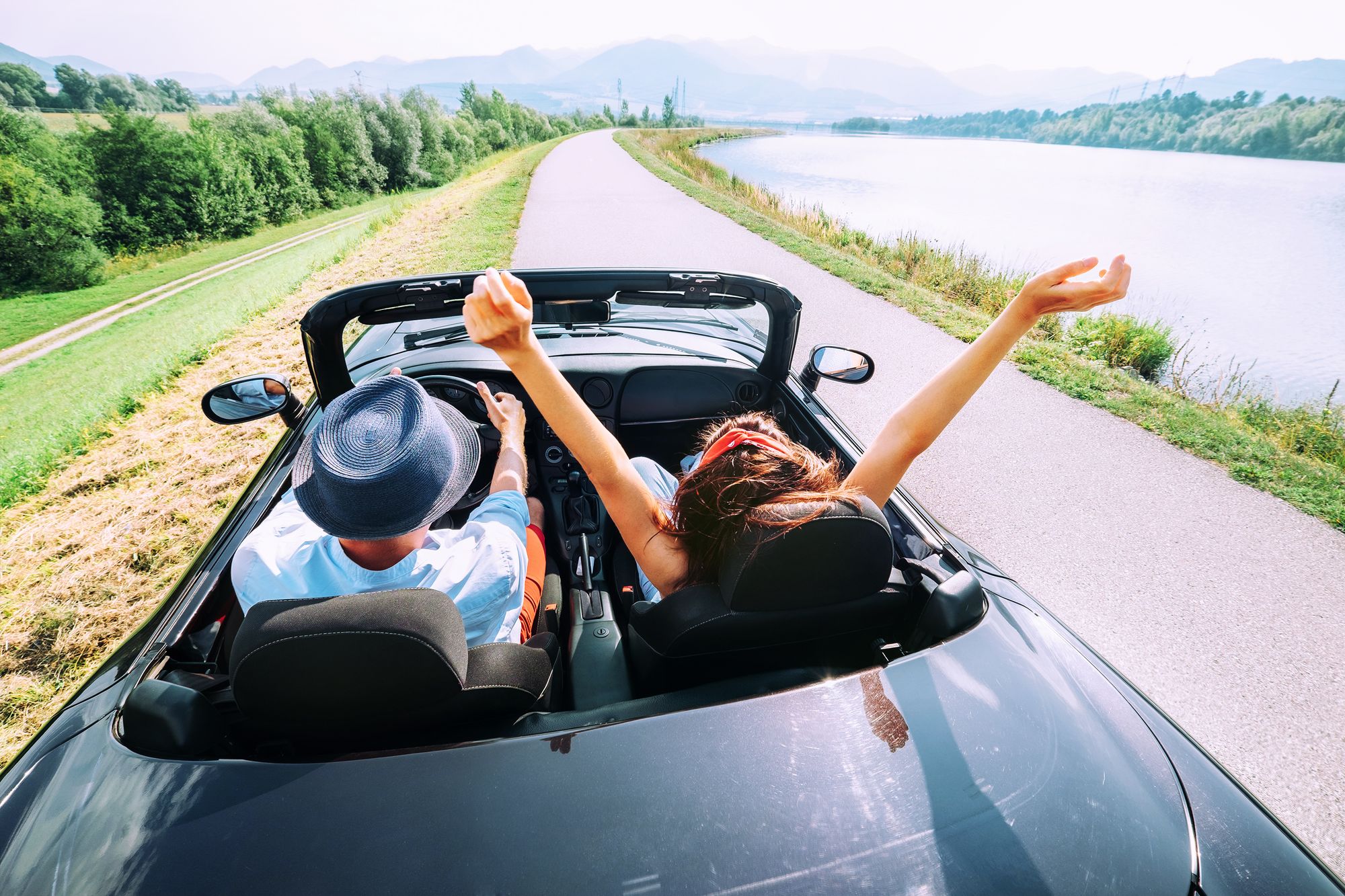 Two people ride in an open convertible, one person throws their hands up