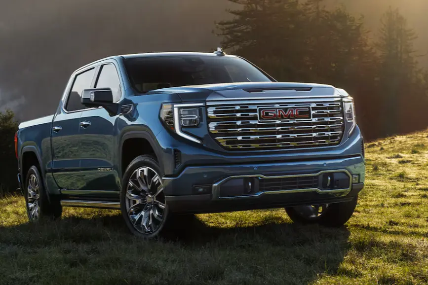 A premium deep blue 2026 GMC Sierra 1500 Denali featuring a chrome grille and multi-spoke luxury wheels parked on a grassy hill at sunrise.