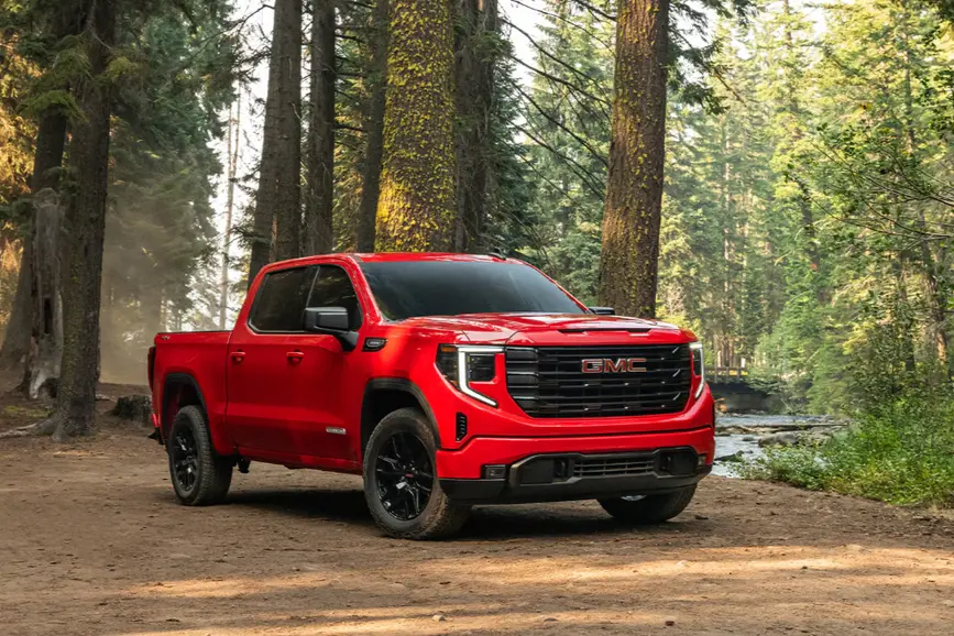 A bright red 2026 GMC Sierra 1500 crew cab truck with black wheels parked on a dirt trail surrounded by tall pine trees in a forest setting.
