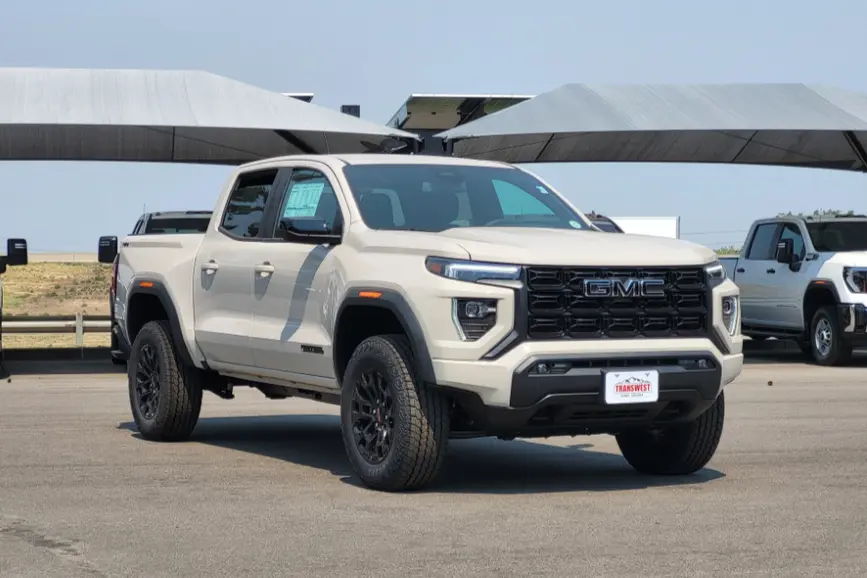Front three-quarter view of a sand-colored 2026 GMC Canyon Elevation mid-size truck with a black grille and black wheels parked on a dealership lot.