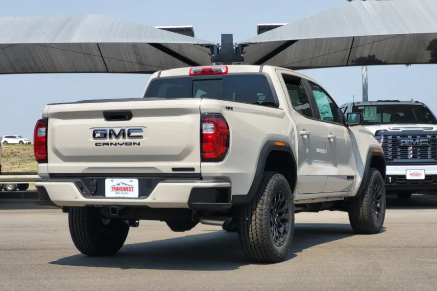 Rear view of a sand-colored 2026 GMC Canyon Elevation truck highlighting the stamped GMC Canyon tailgate and rugged all-terrain tires.