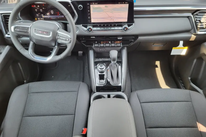 Interior view of a 2026 GMC Canyon Elevation cockpit featuring black cloth front bucket seats, a multi-function steering wheel, and a center infotainment screen showing a navigation map.