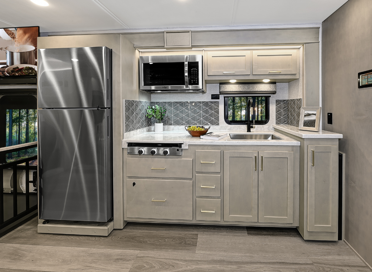 2026 Renegade Valencia RV kitchen with a stainless steel refrigerator, microwave, stovetop, sink, and light wood cabinetry with brass handles, accented by a geometric tile backsplash