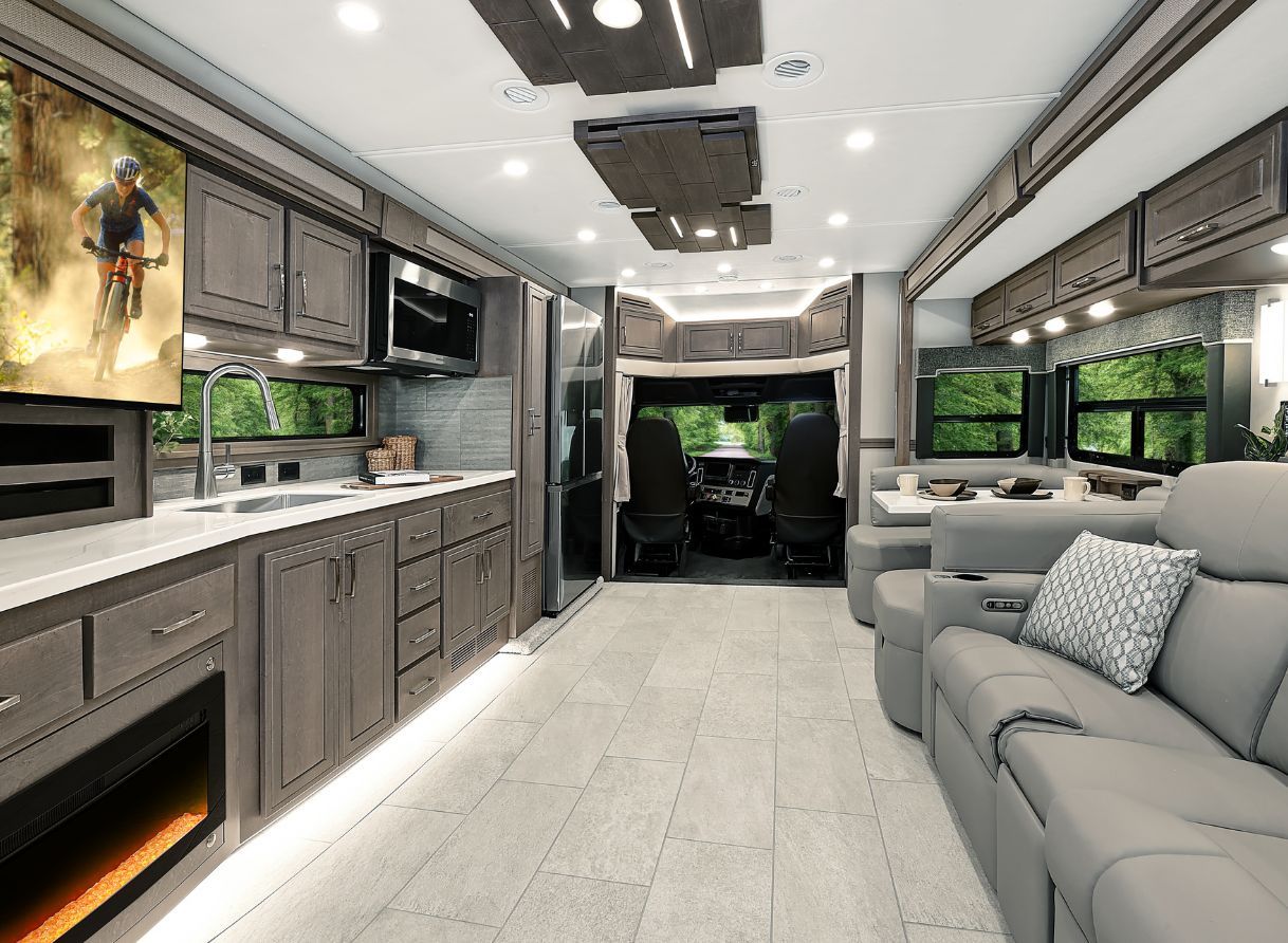 Interior of the 2026 Renegade Explorer looking from the back to the cab, showing the kitchen, entertainment center, and lounge seating. Bright overhead lighting highlights modern cabinetry and open layout.