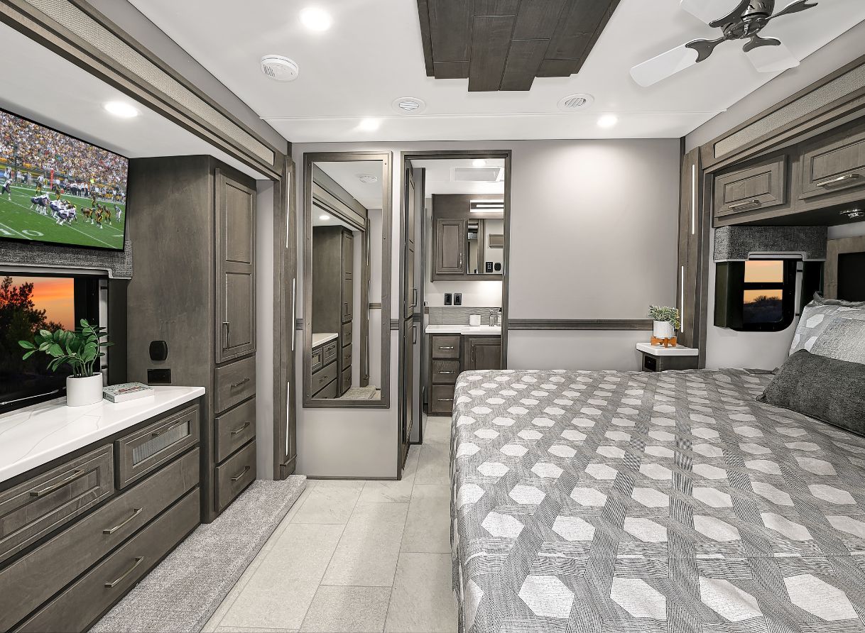 Bedroom inside the 2026 Renegade Explorer with a large bed, overhead cabinets, dresser, and mirrored wardrobe. A mounted TV and windows bring entertainment and natural light.