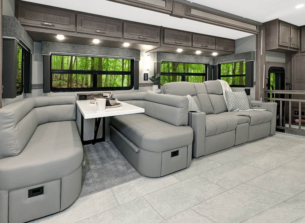 Living area of the 2026 Renegade Explorer with a U-shaped dinette and reclining sofa. Large windows provide forest views and overhead storage runs the length of the wall.