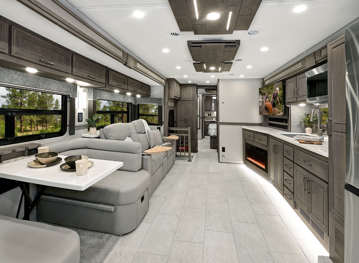 Interior view of the 2026 Renegade Explorer looking toward the bedroom, with a kitchen, fireplace, and lounge seating in the foreground. TV and overhead storage frame the space.