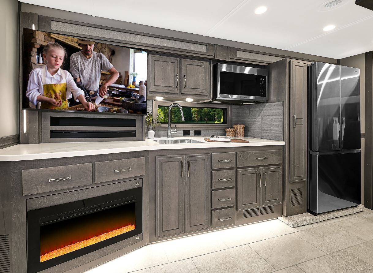 Kitchen inside the 2026 Renegade Explorer with dark cabinetry, stainless steel refrigerator, and microwave. A flat-screen TV sits above the fireplace for entertainment.