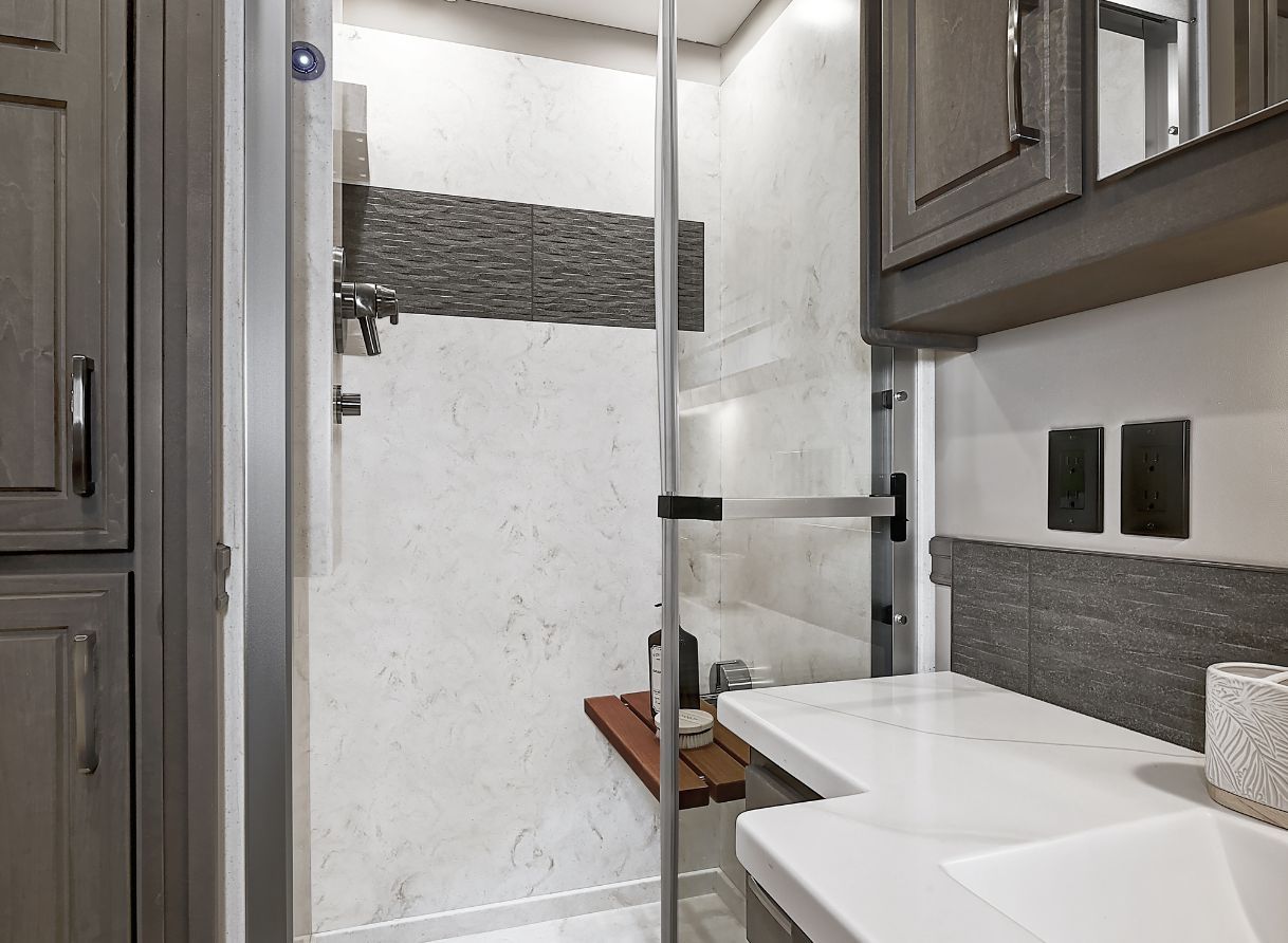 Walk-in shower inside the 2026 Renegade Explorer featuring a glass door, wall accents, and a fold-down wooden seat. Modern tile finishes complete the look.
