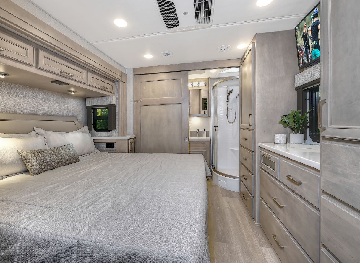 2026 Renegade Verona master bedroom with king bed, overhead storage, dresser, TV, and en-suite bath.