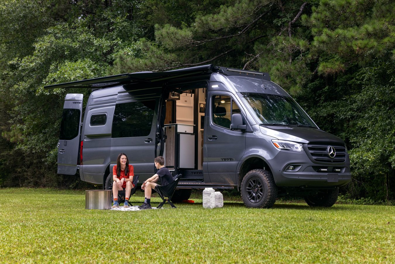 A child and an adult sit outside a 2026 Tiffin GH2 on a sunny day