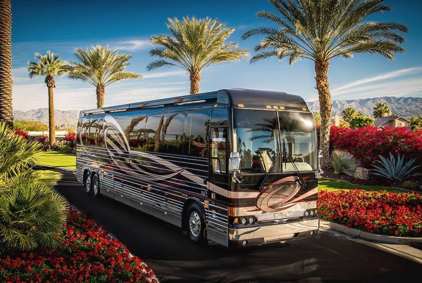 An Emerald Luxury Coach RV driving along a road lined with flowerbeds and palm trees