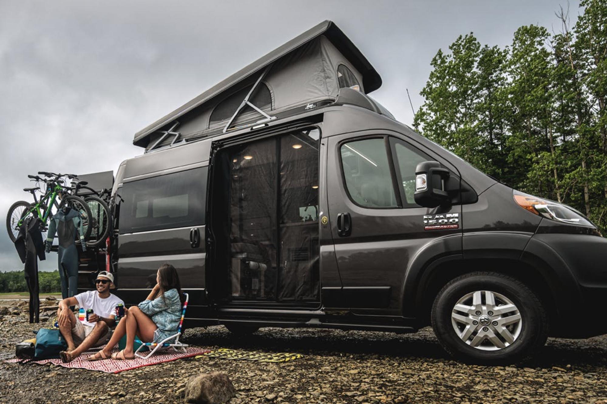 Black Thor campervan parked on a rocky beach with a young couple sitting on a blanket in front of it