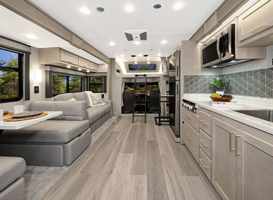 Interior of a 2026 Renegade Valencia RV looking toward the cab, featuring a sofa, booth dinette, full kitchen with stainless steel appliances, and a ladder leading to the overhead bunk above the driver’s area.