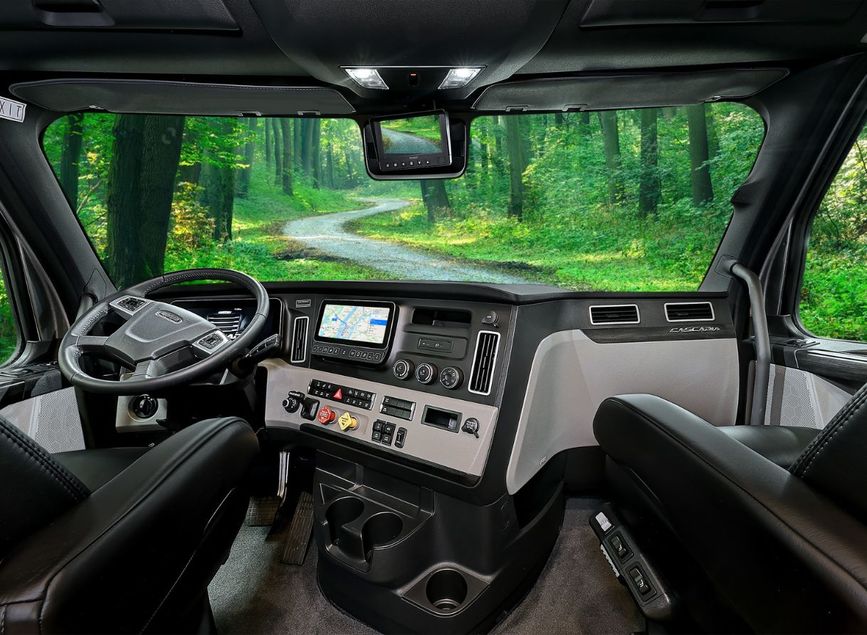 Driver’s cab of the 2026 Renegade Explorer with leather seating, touchscreen navigation, and modern controls. Large windshield view shows a wooded trail winding through the forest.