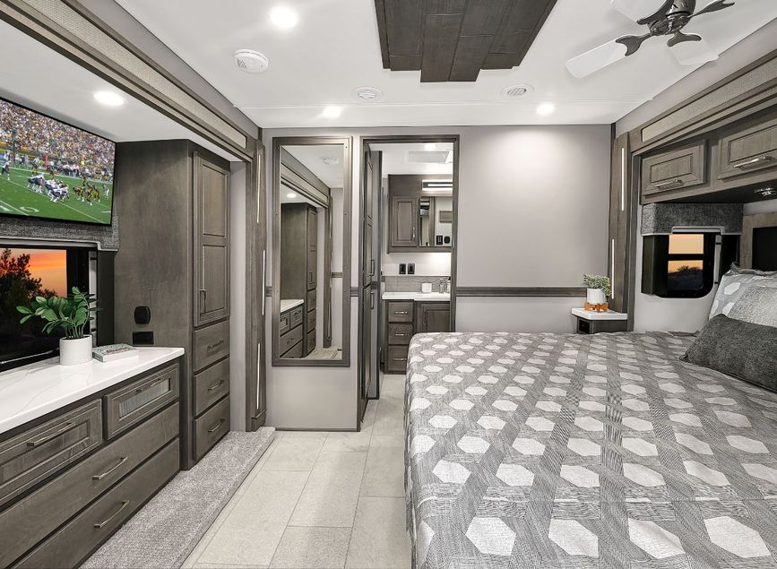 Bedroom of the 2026 Renegade Explorer with a large bed, overhead storage, and built-in dresser with TV. A mirrored wardrobe leads into the bathroom, adding light and space to the room.