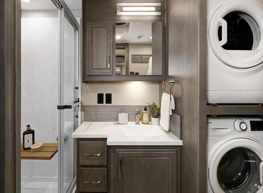 Bathroom in the 2026 Renegade Explorer with vanity, storage cabinets, and a glass-door shower. A stacked washer and dryer sit beside the sink for added convenience.
