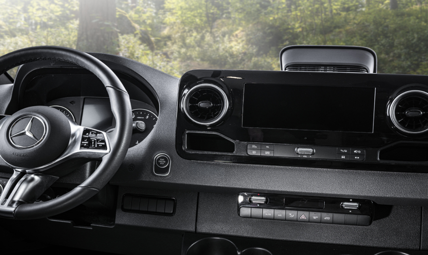 Mercedes-Benz Sprinter cockpit inside a Renegade Villagio RV, featuring a leather-wrapped steering wheel, digital display cluster, central infotainment screen, and climate controls with a forest view through the windshield