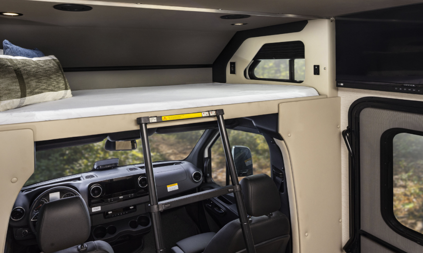Interior of a Renegade Villagio RV showing an over-cab sleeping area with a mattress, ladder access, built-in lighting, and a front view down into the Mercedes-Benz Sprinter cockpit below