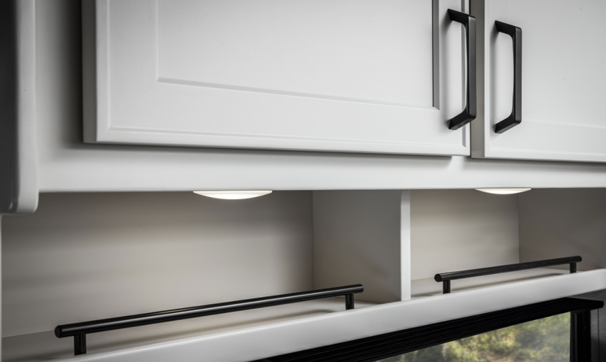 Interior detail of a Renegade Villagio RV showing upper kitchen cabinetry with white shaker-style doors, black hardware, and built-in under-cabinet lighting above open storage shelves