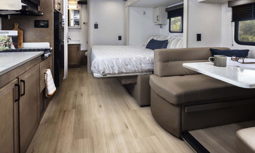 Interior of a Renegade Villagio RV showing a compact kitchen with cooktop and cabinetry, a booth dinette with table, and a slide-out bed in the back with light wood flooring and neutral finishes