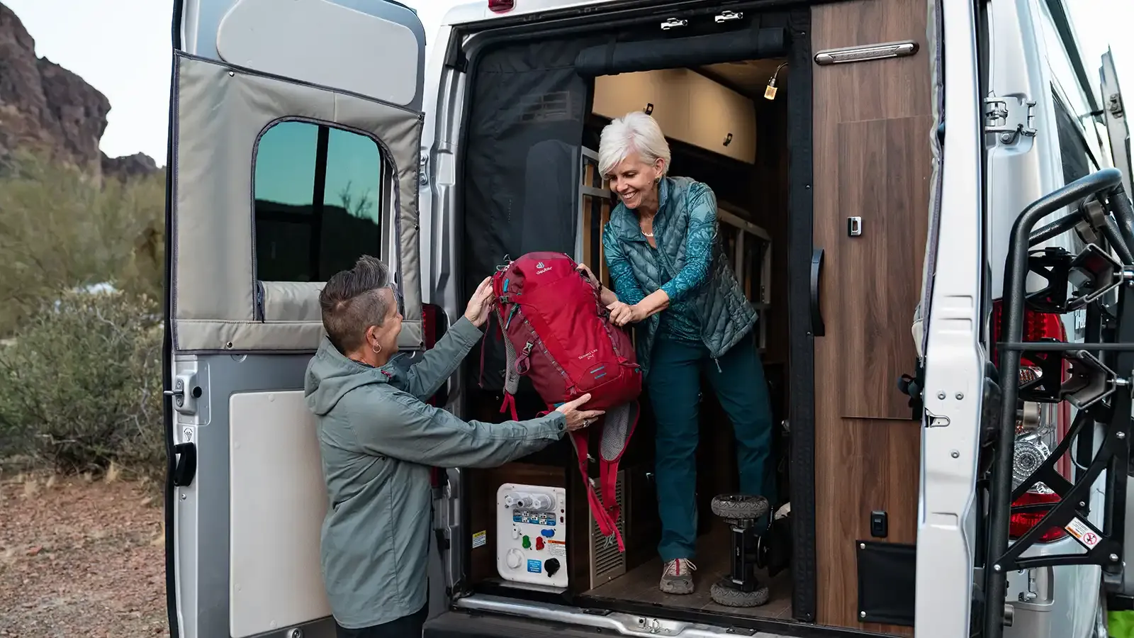 Two people unload a backpack from the back of a Travato